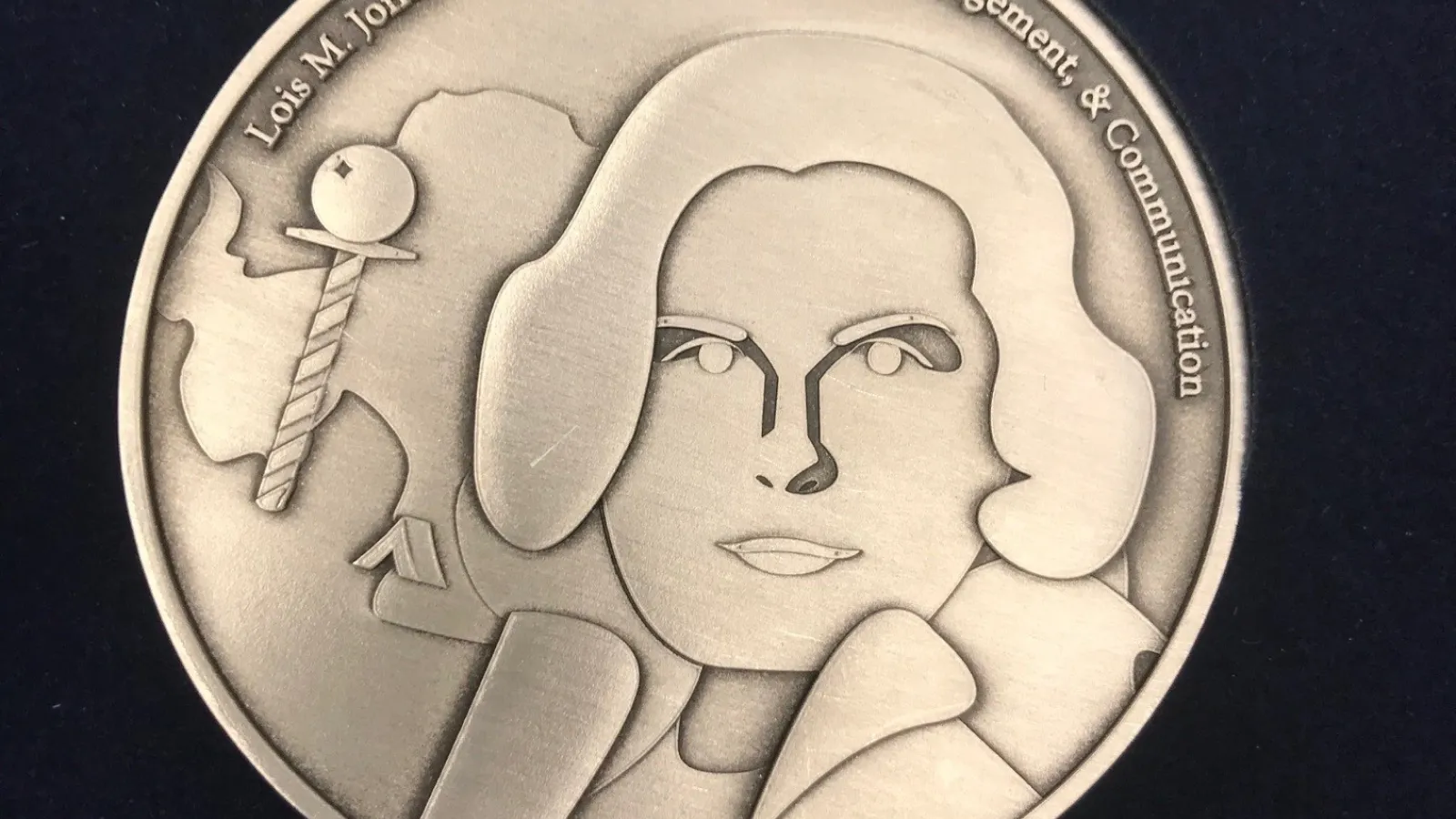 Lois Jones Medal Front. It is a gold medalion engraved with the head and shoulder of a woman with a bob haircut and a coat. Beside her is an aerial view of the Antarctican continent. On the top lip of the medalion, the full name of the award has been engraved.