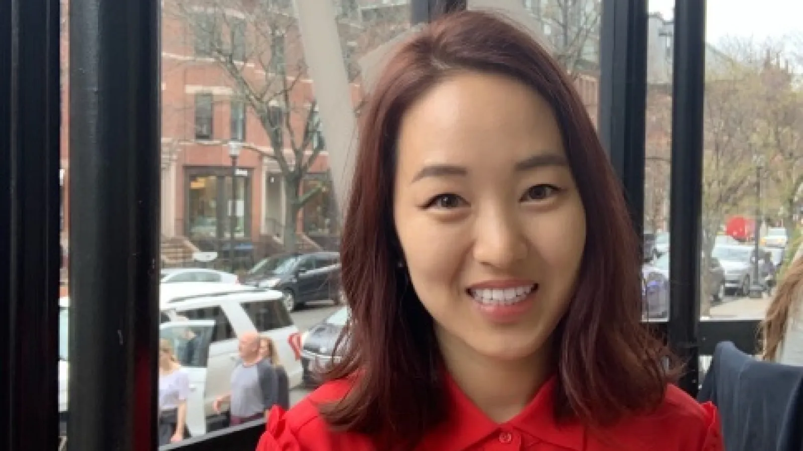 Smiling image of Kyungmin Sung with long sleeve red button shirt and shoulder length reddish straight hair inside in front of a series of windows with parked cars in background.