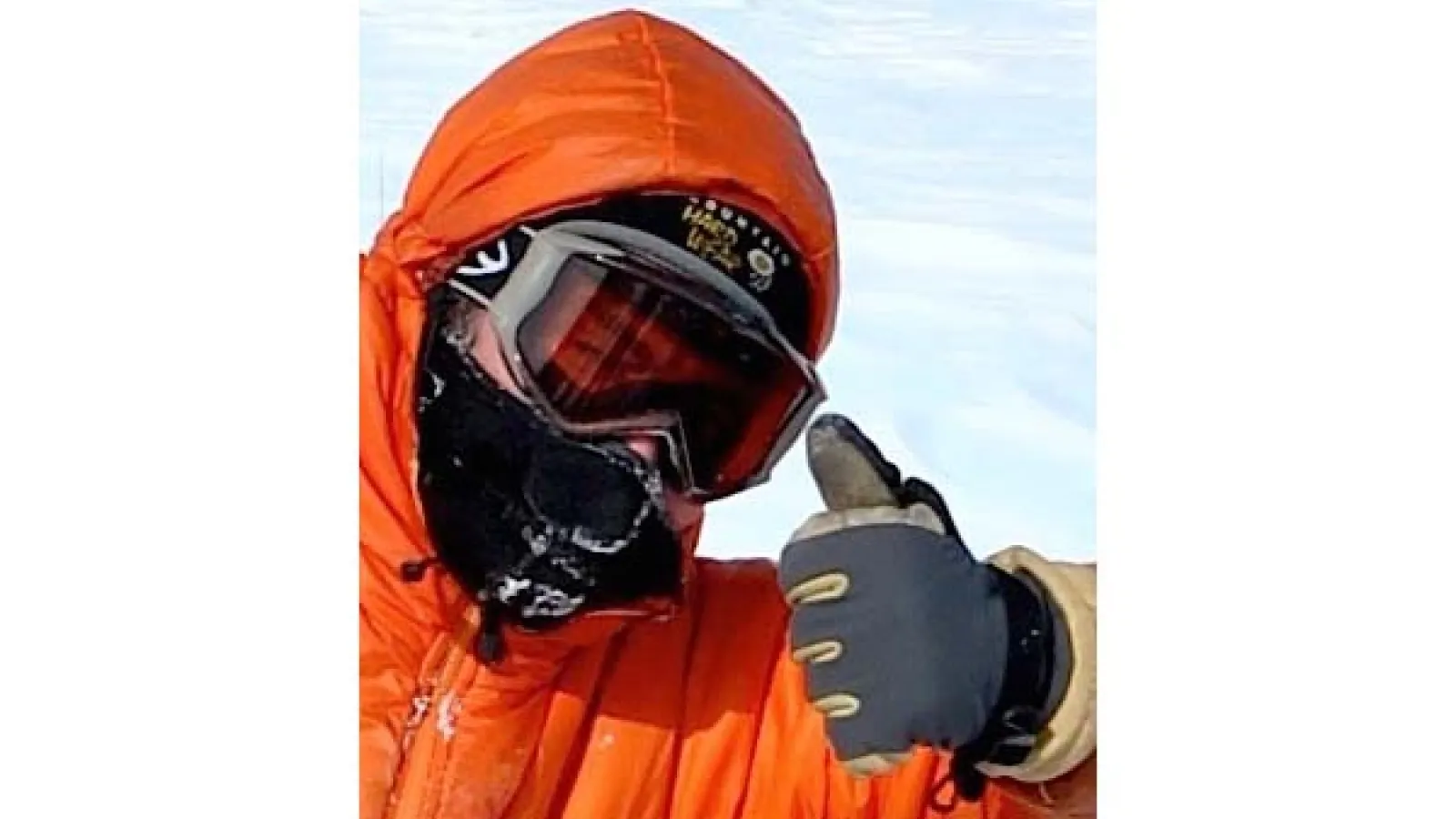 head and left hand and arm shot of Ian Howat wearing and orange buff jacket with a hood and wearing orange lens googles with gray frame and black mask covering mouth and nose giving a thumbs up  wearing a black and tan glove with white clouds covering blue sky in background