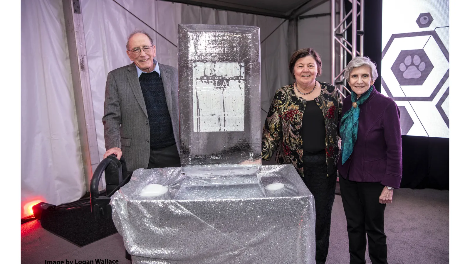 Left to right: Lonnie Thompson, Georgie Shockey and Ellen Mosley-Thompson standing in a room around a table with silver table cloth and a rectangular ice sculpture  on it with lilac colored carpet and white background with metal wires, and scaffolding