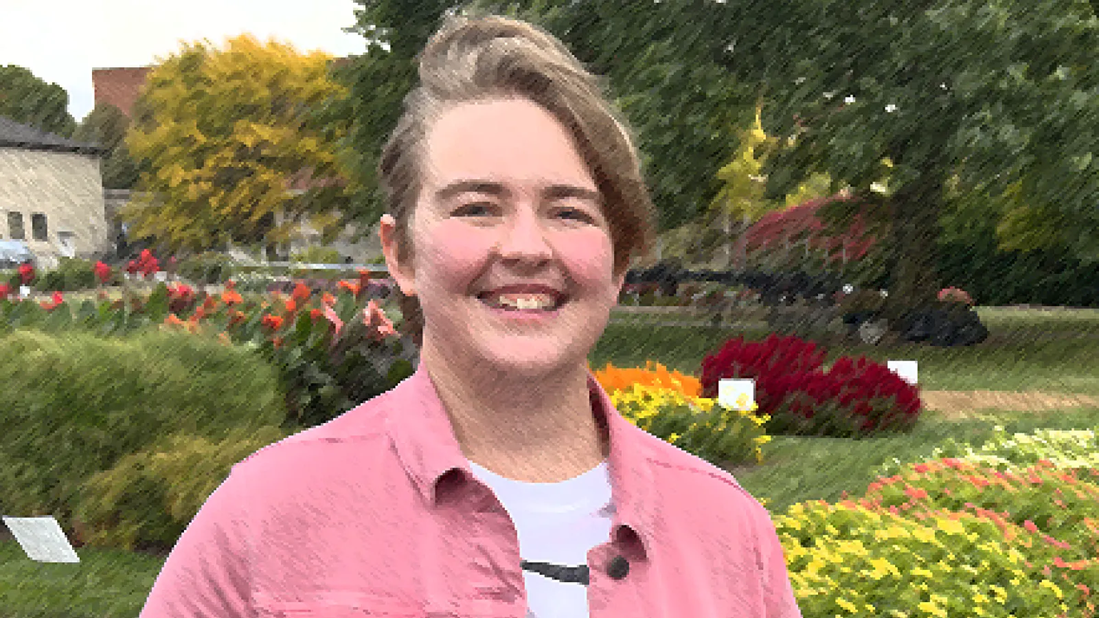 Standing in a garden is a white woman with short blonde hair wearing a pink jacket