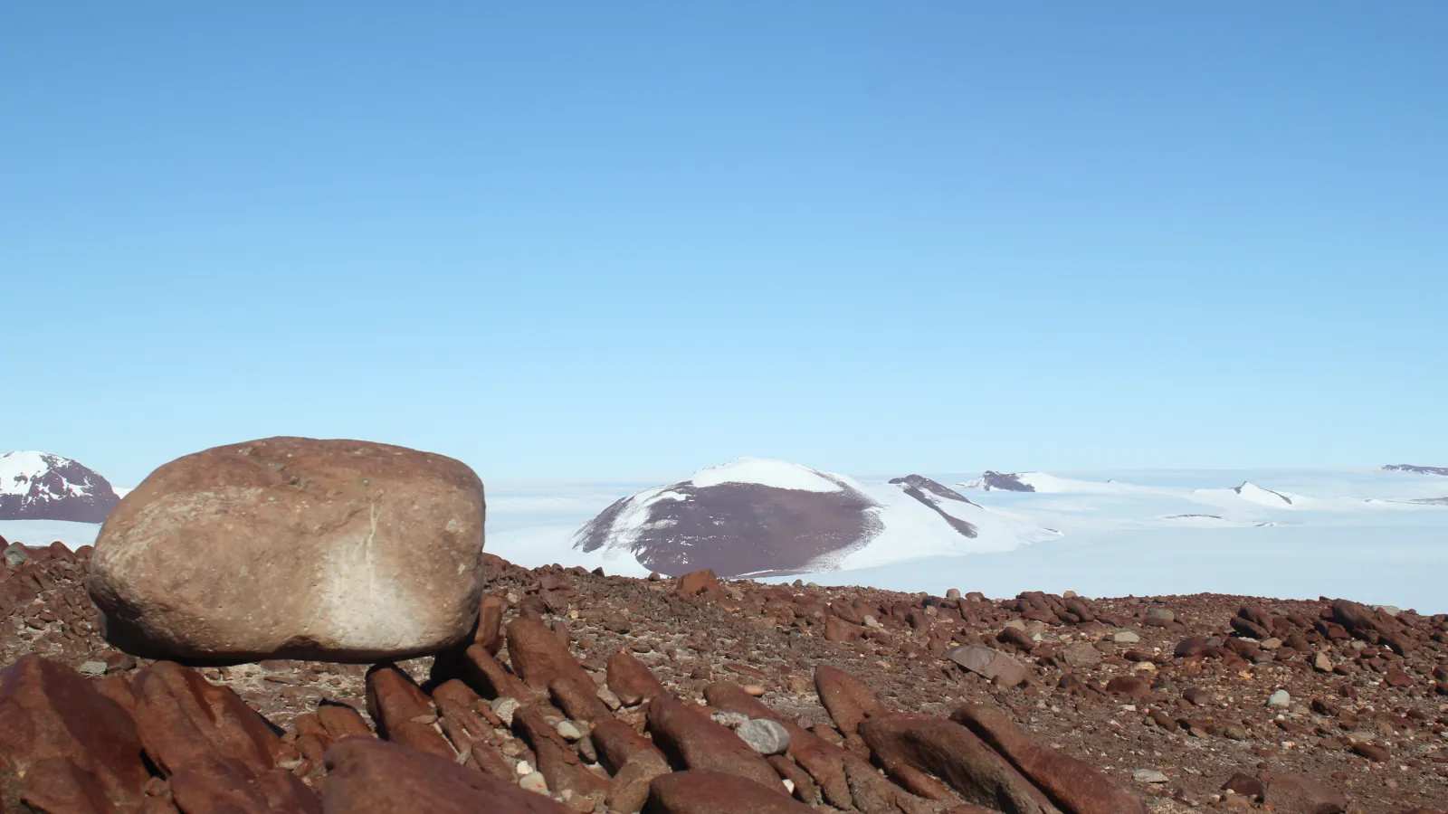 a brown rocky ground with a boulder in the foreground and snow covered landscape in the background.