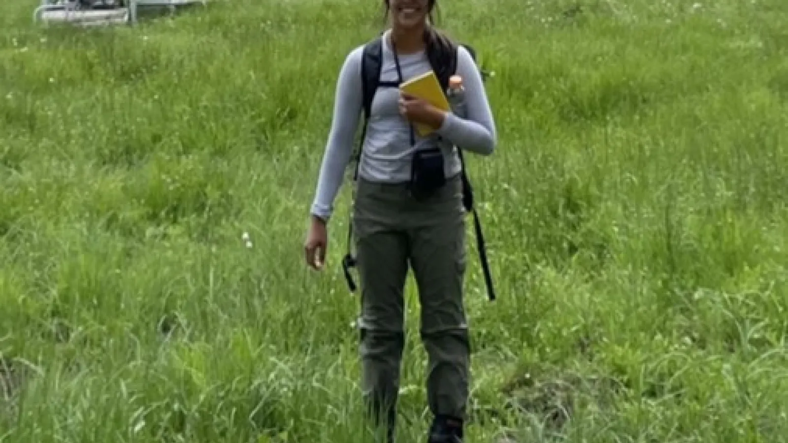 a person in a green grass field with notebook, backpack, ball cap smiling at the camera.