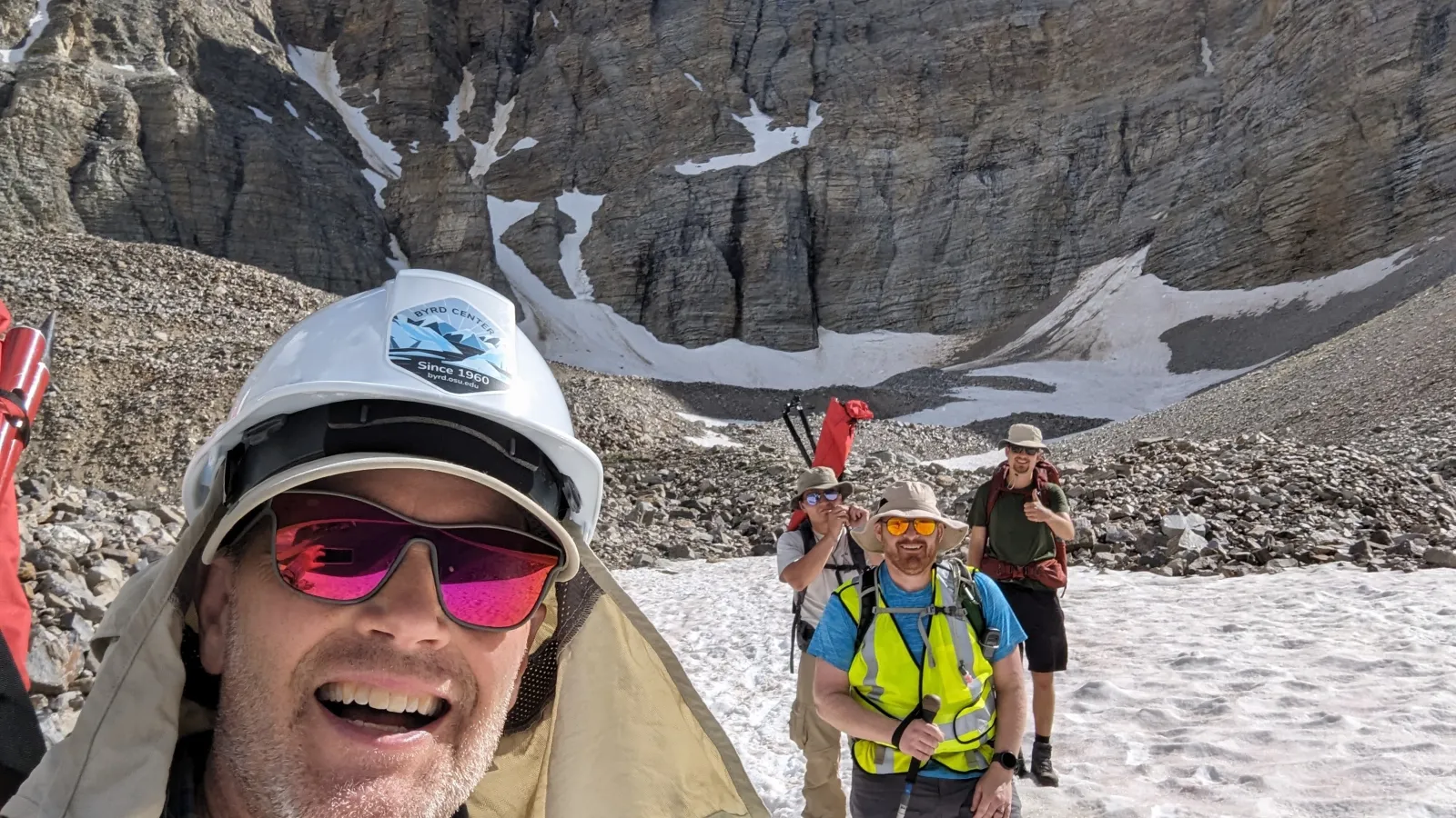 Four people smiling, posing surrounded by rocky mountains and snow.