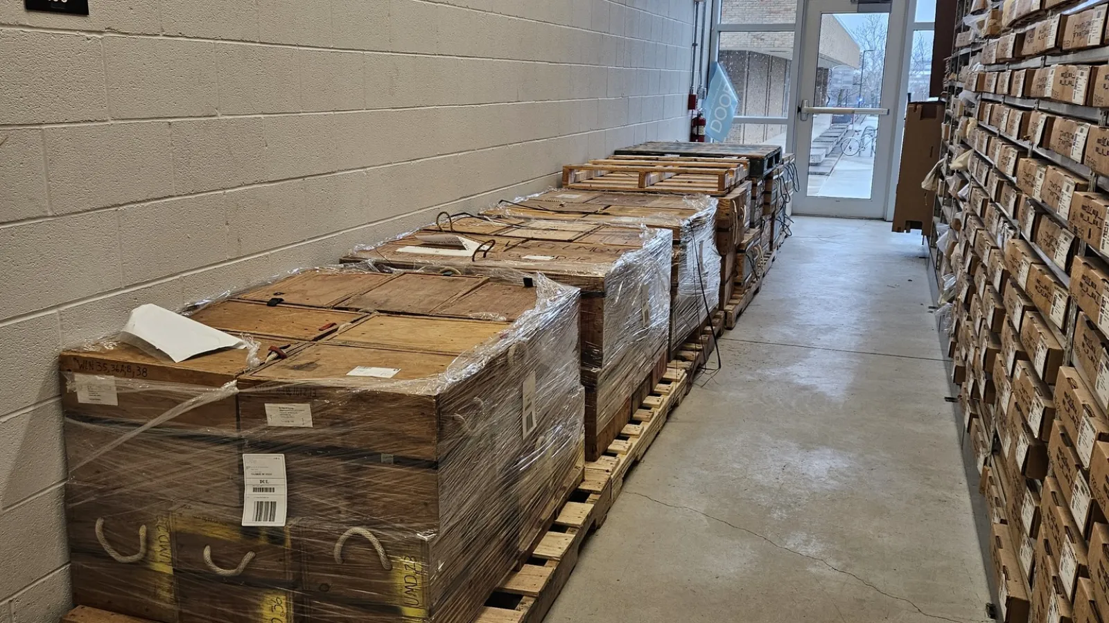 Pallets of wooden boxes in a row along the wall with shelving of boxes on the opposite side and a glass window and door at the end of the isle.