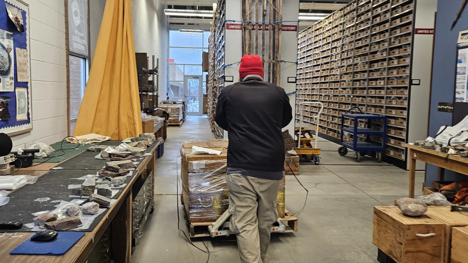 A man pushing a cart with a pallet of wooden boxes inside a warehouse with shelves filled with boxes.