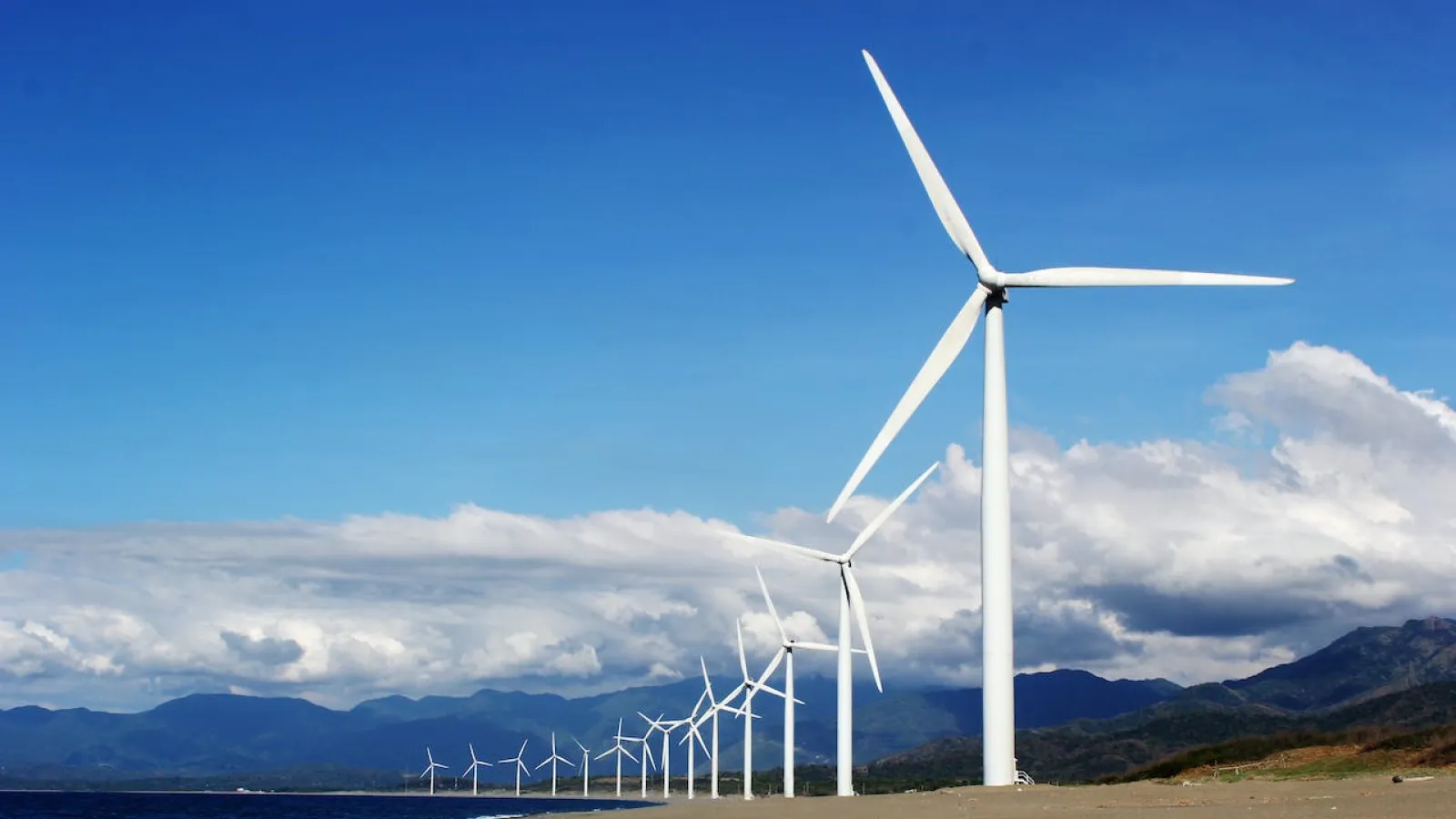 White wind turbines on sand near body of water under blue skies with white clouds.