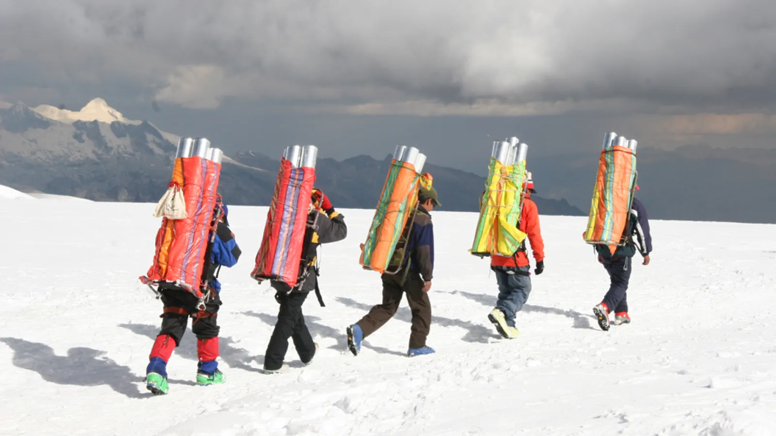 Five people, each carrying a group of long cylindrical tubes on their backs, walking away in a line on snow covered ground with snow capped mountain range at a difference under gray cloudy sky.