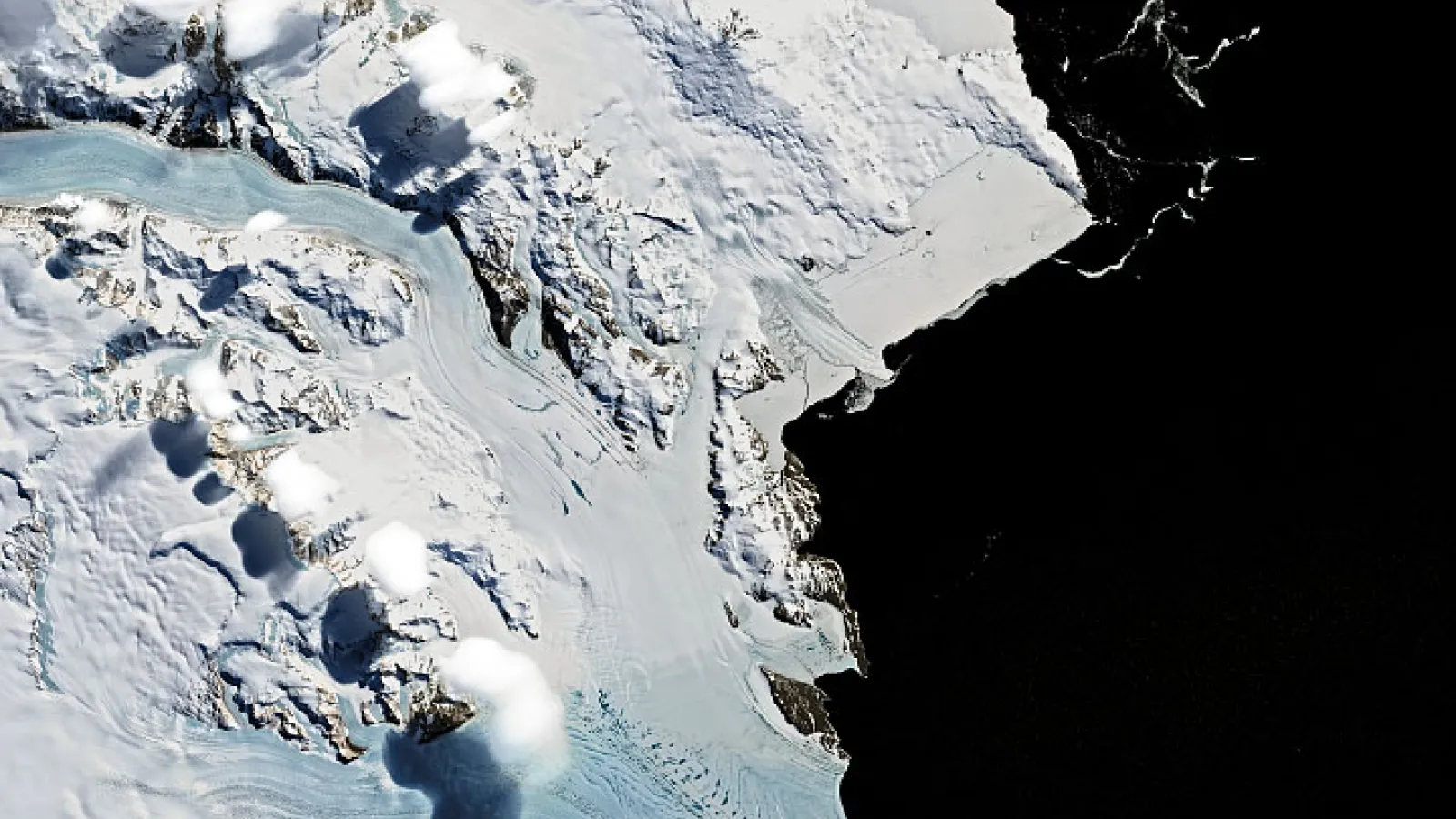 Aerial view of glaciers, snow-covered mountains and white, puffy clouds against a black background. 