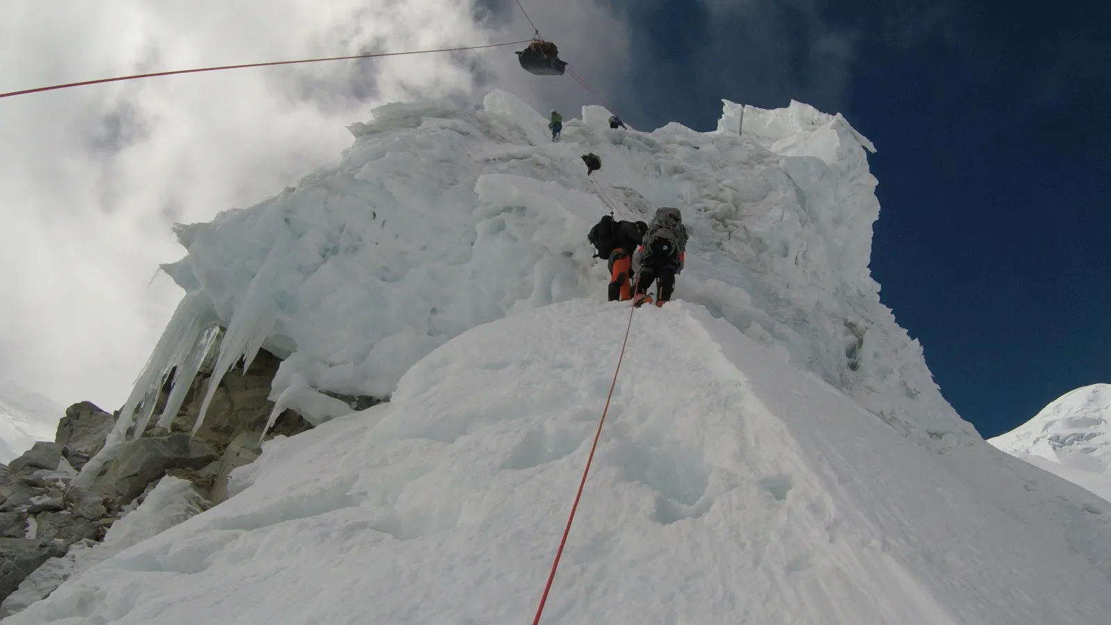 Researchers ascending a steep, icy section of a mountain and moving equipment, using fixed ropes for safety.