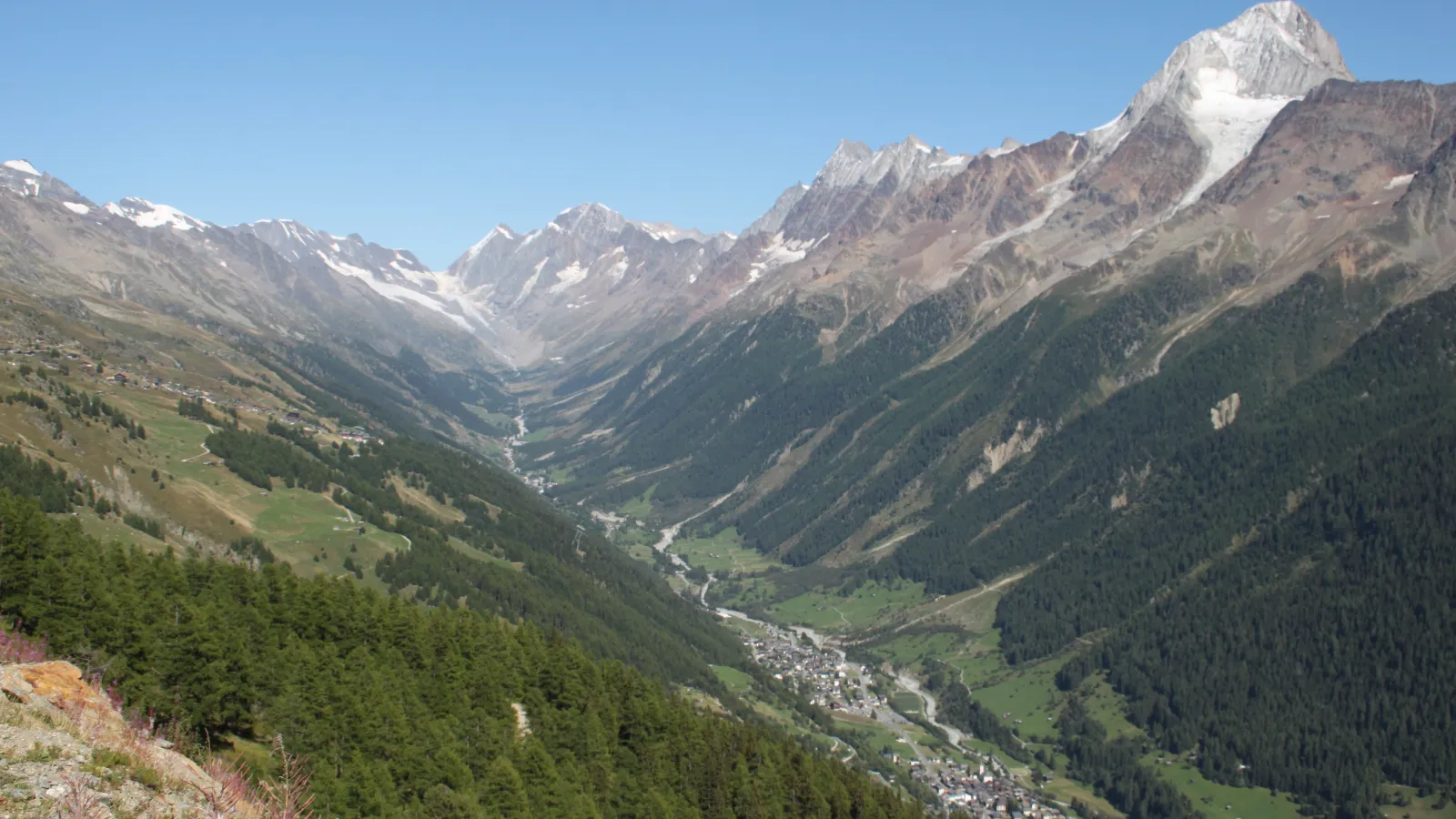 A wide valley with alpine villages in the middle surrounded by steep forested slopes and high mountain peaks with glaciers.