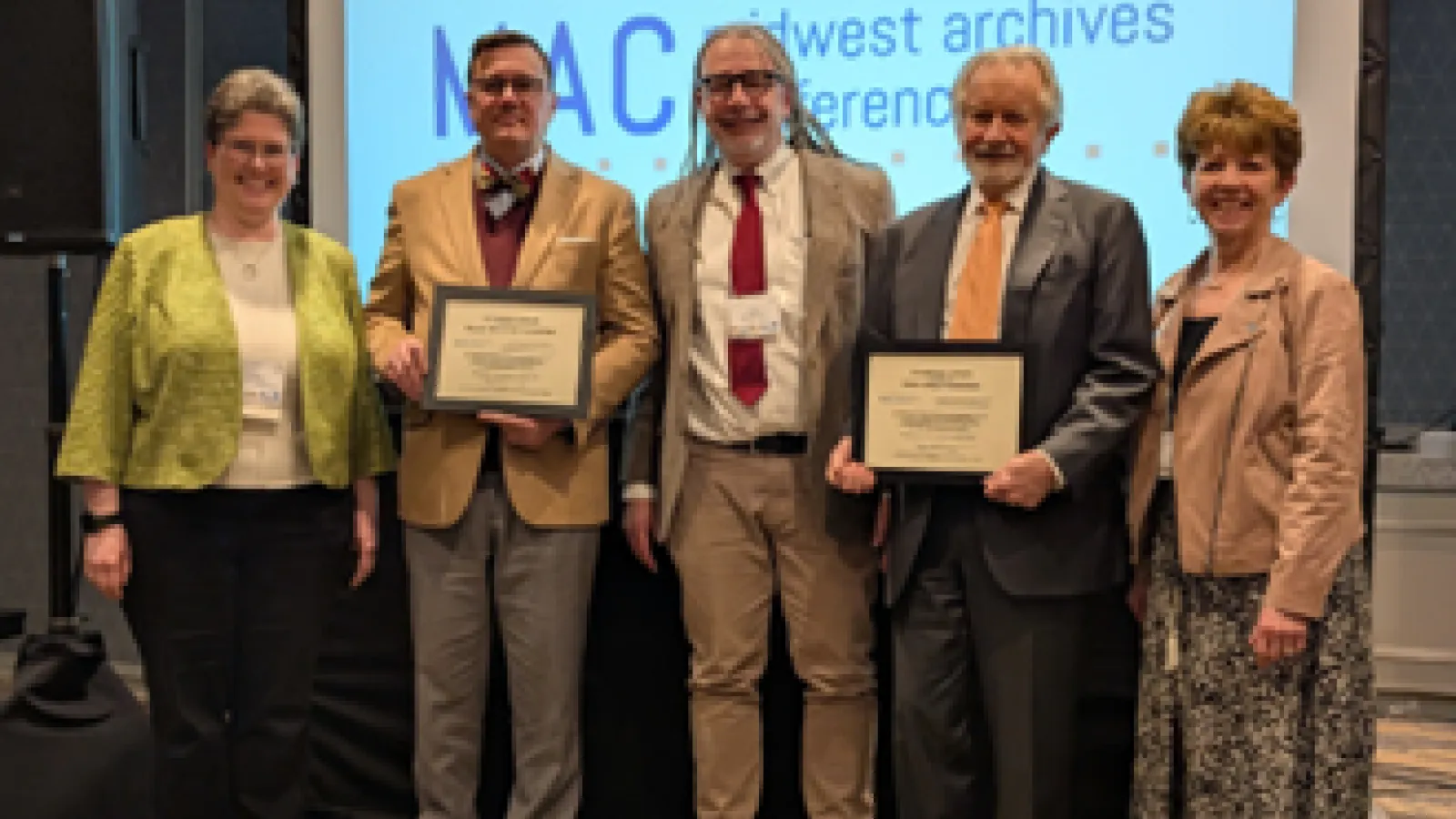 Five individuals, two holding certificates, stand in front of a presentation screen displaying "MAC Midwest Archives Conference".