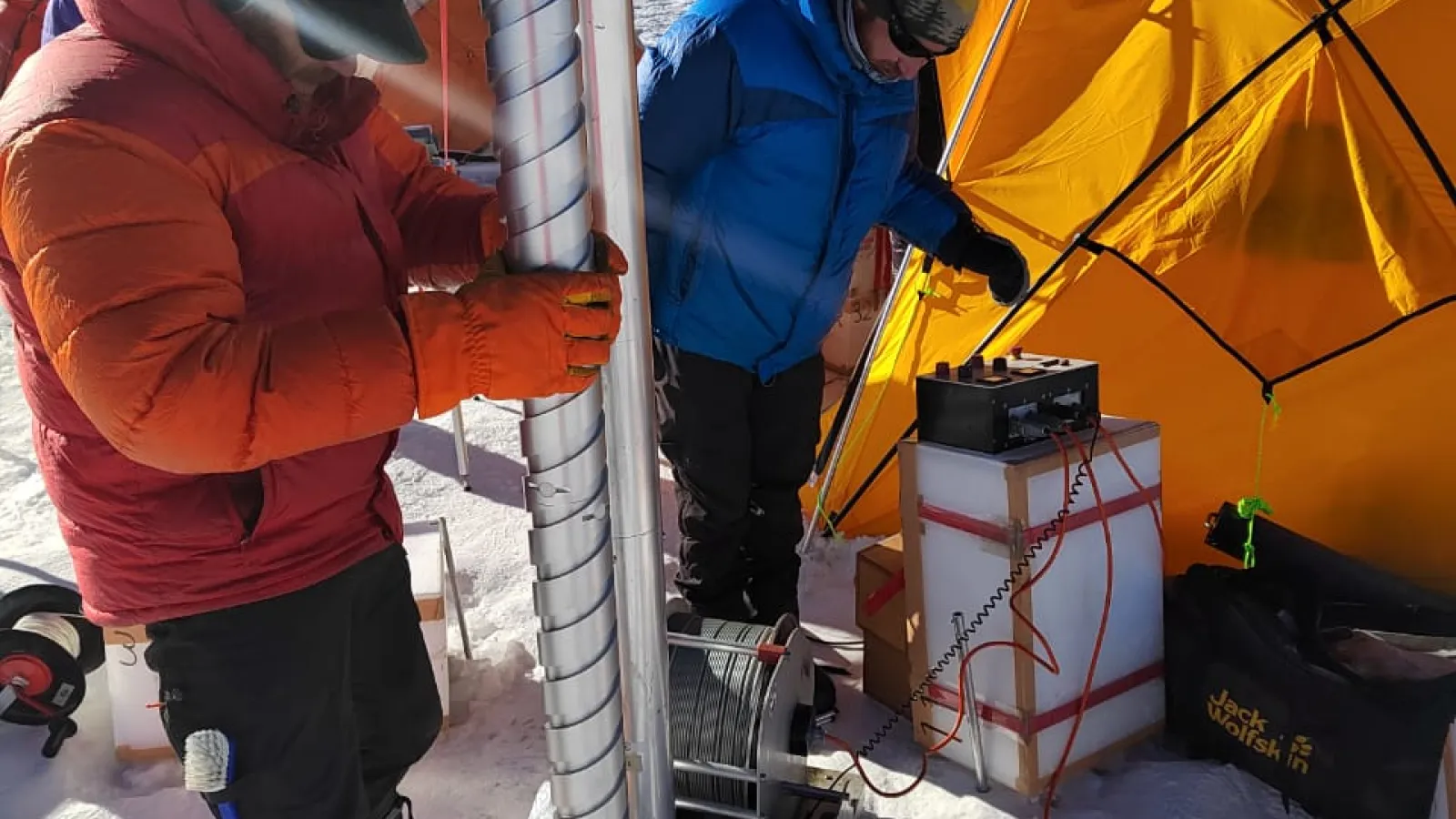 Two people are setting up a tall drill Ice core beside an orange dome tent.