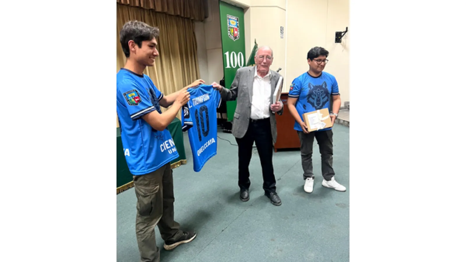 Two students in blue jerseys present a personalized soccer jersey and gift to a guest in a gray blazer at the front of an auditorium.