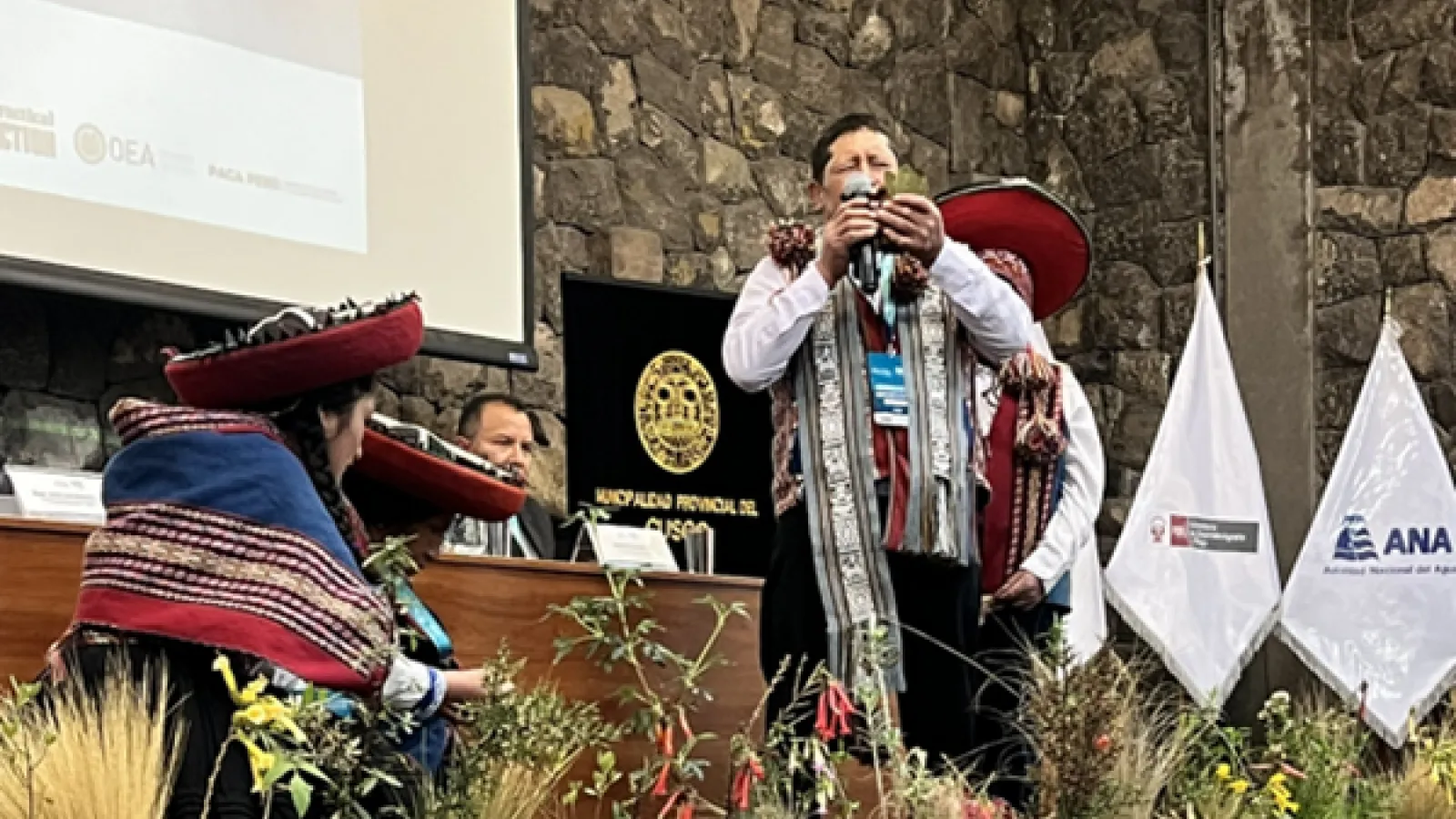 indigenous ceremony in front of  a stone-walled stage with plants in front, as a man in traditional attire holds an offering