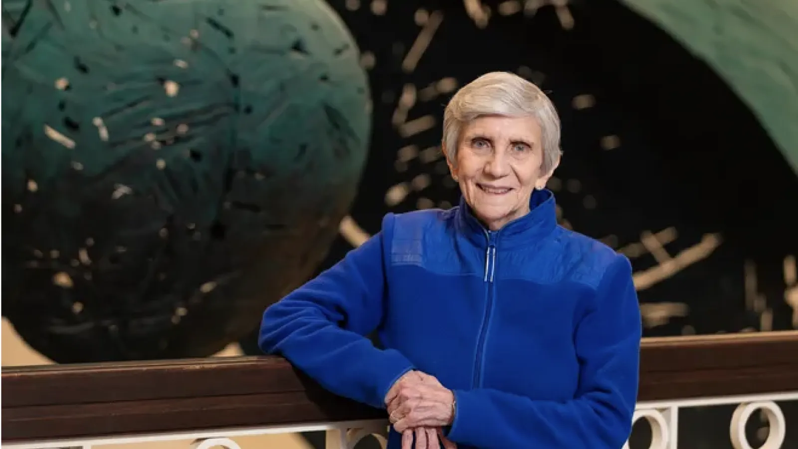 Ellen Mosely-Thompson wearing a blue zip-up jacket smiling and leaning against a railing. 