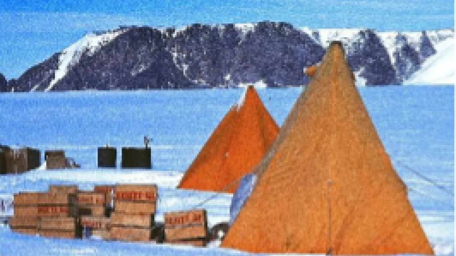 Polar rock boxes sitting in the snow next to a large orange tent with mountains in the background. 