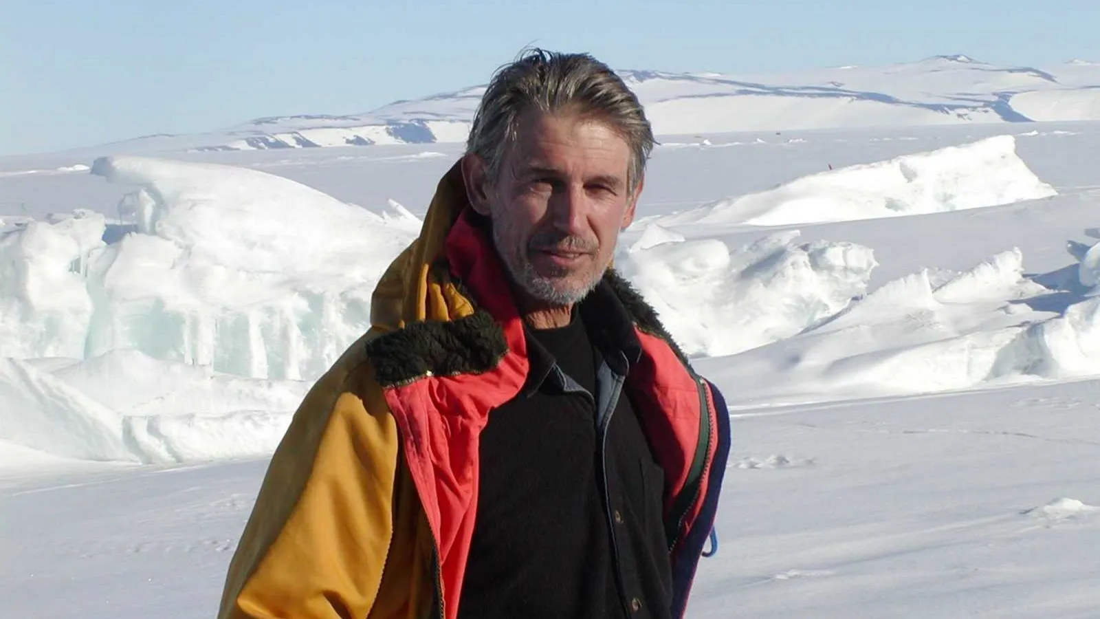 Peter Barrett in a red and yellow cold-weather jacket standing on an Antarctic ice shelf, surrounded by snow and ice formations with distant mountains in the background.