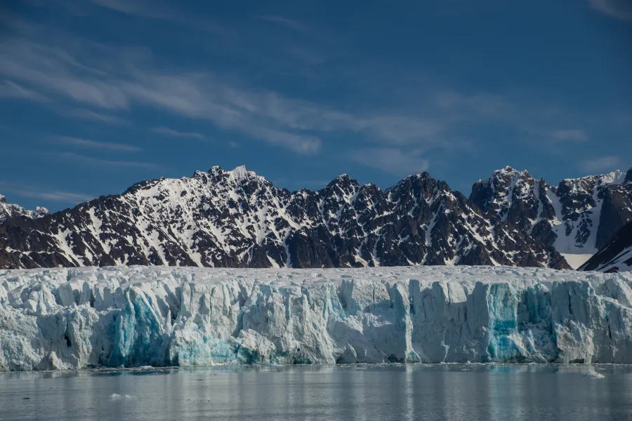 A pano of a lake and glacier and a rocky mountain with snow streaks under blue skies and light, white clouds