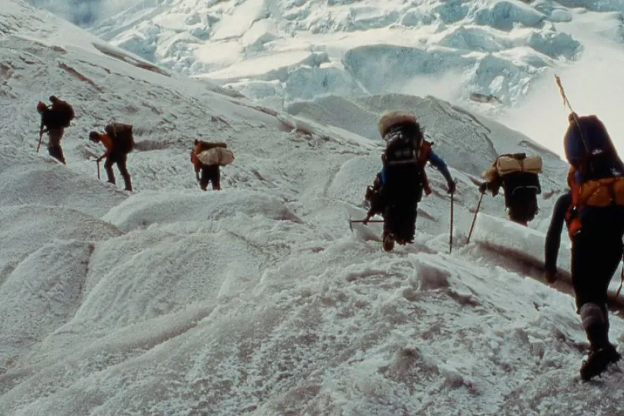 Climbers hike up a snowy glacier ridge with ice cliffs behind them.