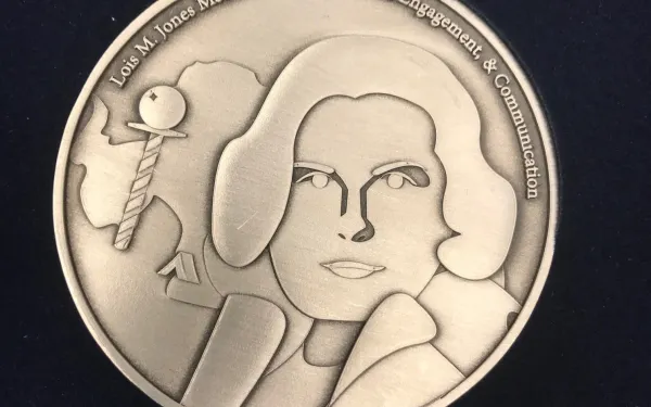 Lois Jones Medal Front. It is a gold medalion engraved with the head and shoulder of a woman with a bob haircut and a coat. Beside her is an aerial view of the Antarctican continent. On the top lip of the medalion, the full name of the award has been engraved.