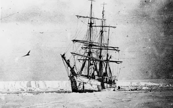 Black and white photo of sail ship with sails down in snow with bird flying close by and light gray skies 