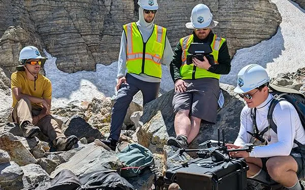 A group standing and sitting on rocks in a snow covered mountainous  regions wearing yellow vests and hard hats working on a drone.