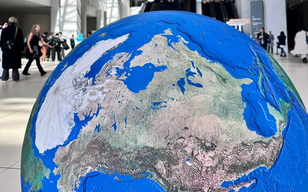 A large, detailed globe model  positioned in the foreground of a spacious convention center lobby. People walk and gather in the background near escalators.