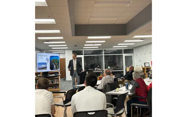 A presentation with an audience listening to a speaker standing next to a screen.