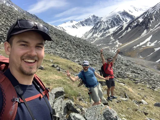 Three people in a rocky field smiling for the camera. In a distance there is a snow covered mountain.