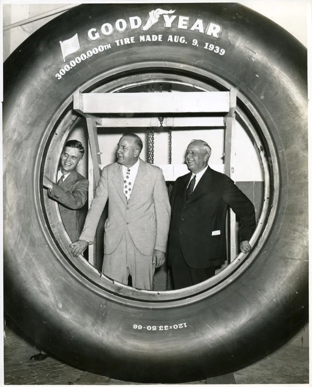 Three men in suits standing behind a very large Goodyear tire, which frames them. Text on the tire reads "300,000,000th tire made August 9, 1939."