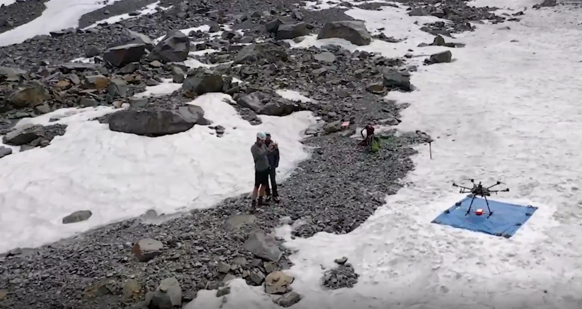 Three individuals conducting research or fieldwork on a rocky glacier area, with a drone set up nearby.