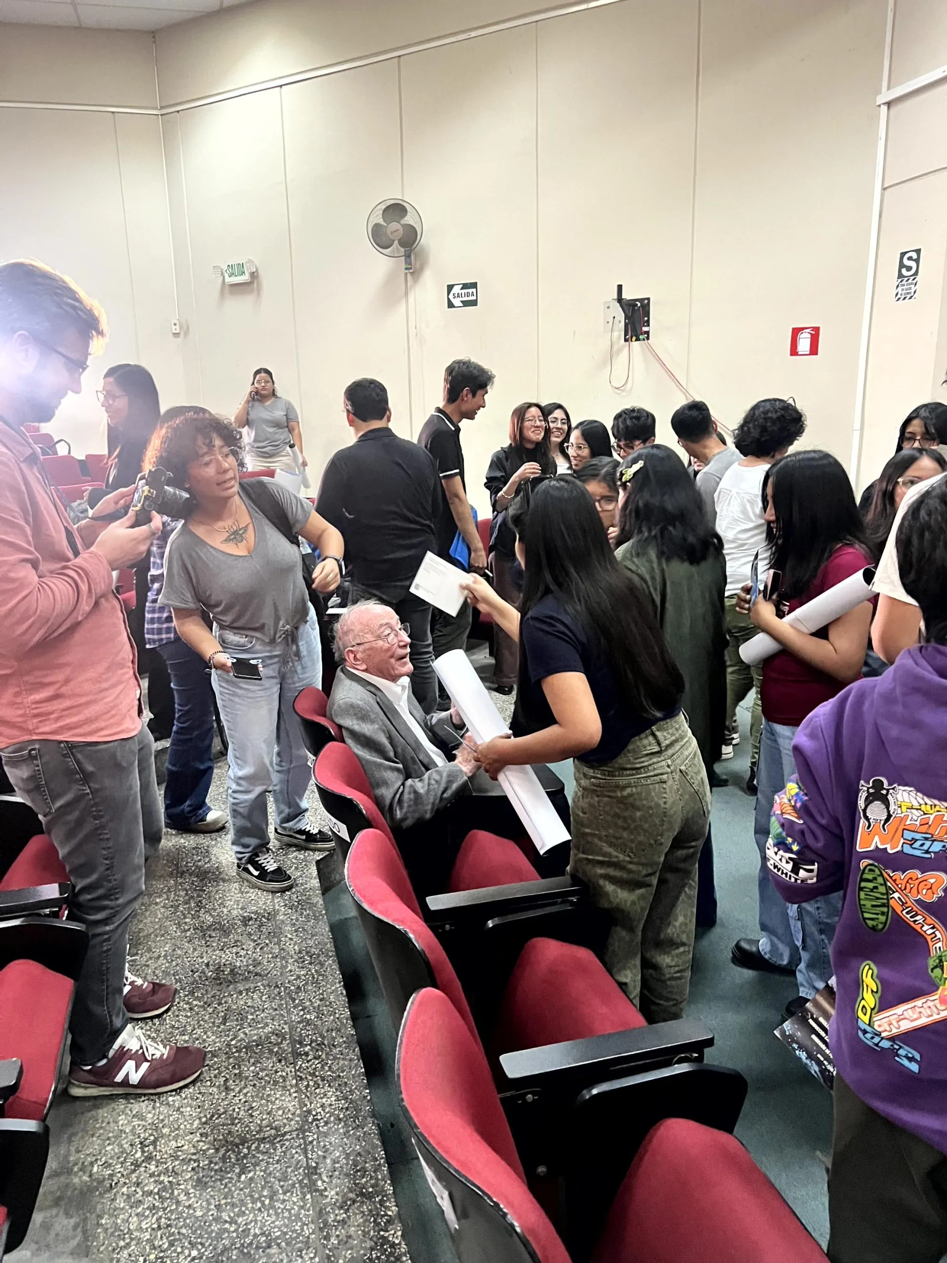 people gather around an older man seated in an auditorium, chatting and holding papers after an event.