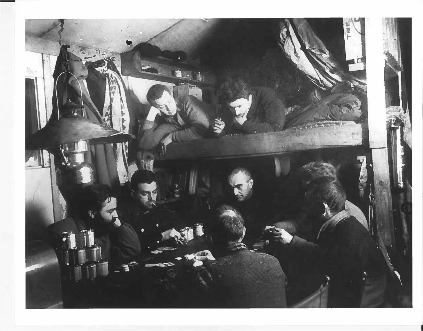 Black-and-white photo of several men playing cards around a table in a cramped bunkroom, while two others lean over an upper bunk to watch.
