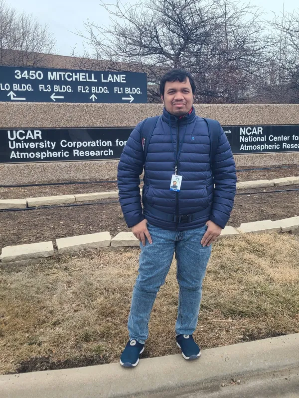 A man in a puffer jacket standing in front of a large outdoor sign. The sign points towards different atmospheric research buildings.