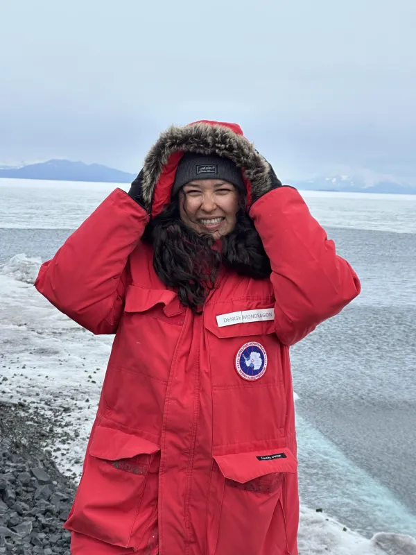 Girl with long black curly hair wearing big red jacket in Antarctica. Hands are on head protecting head from wind. 