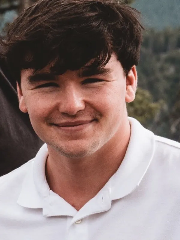 Aiden Fuller wearing a white polo shirt smiling with trees in the distance. 
