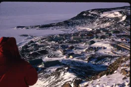 A view of a research facilities and roades from the top of a hill. The terrain is covered in snow. There are epople in red coats moving up the incline.