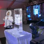 Andrea Bowlin, OSU Advancement, in a white short puff Eskimo jacket standing to the left of an ice sculpture on a table in a room with white walls and red spotlight coming from the ground on the wall